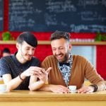 Dois homens estão sentados ao balcão de um café, um sorriso e um olhar juntos para um smartphone. Cada um tem uma chávena de café à sua frente, compartilhando risadas sobre alguns novos livros infantis com temática LGBT que descobri recentemente.