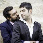 Dois homens de fato, com um a abraçar o outro por trás, estão encostados numa parede de pedra, personificando personalidades gays importantes na história.