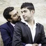 Dois homens de fato, com um a abraçar o outro por trás, estão encostados numa parede de pedra, personificando personalidades gays importantes na história.