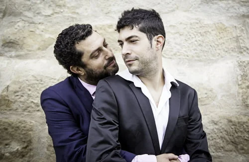 Dois homens de fato, com um a abraçar o outro por trás, estão encostados numa parede de pedra, personificando personalidades gays importantes na história.