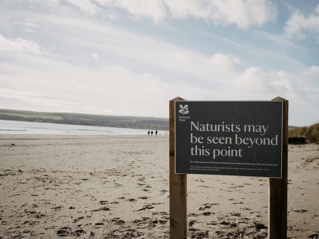 regras praias naturistas Uma placa na praia de areia diz: "Pode haver naturistas para além deste ponto." A praia estende-se até ao horizonte, com algumas pessoas visíveis ao longo. Para os iniciantes a explorar o naturismo, este trecho sereno oferece uma introdução acolhedora.