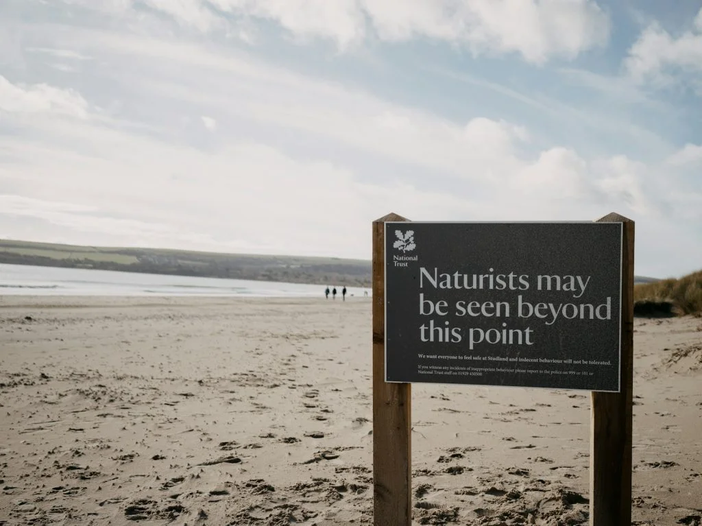 regras praias naturistas Uma placa na praia de areia diz: "Pode haver naturistas para além deste ponto." A praia estende-se até ao horizonte, com algumas pessoas visíveis ao longo. Para os iniciantes a explorar o naturismo, este trecho sereno oferece uma introdução acolhedora.