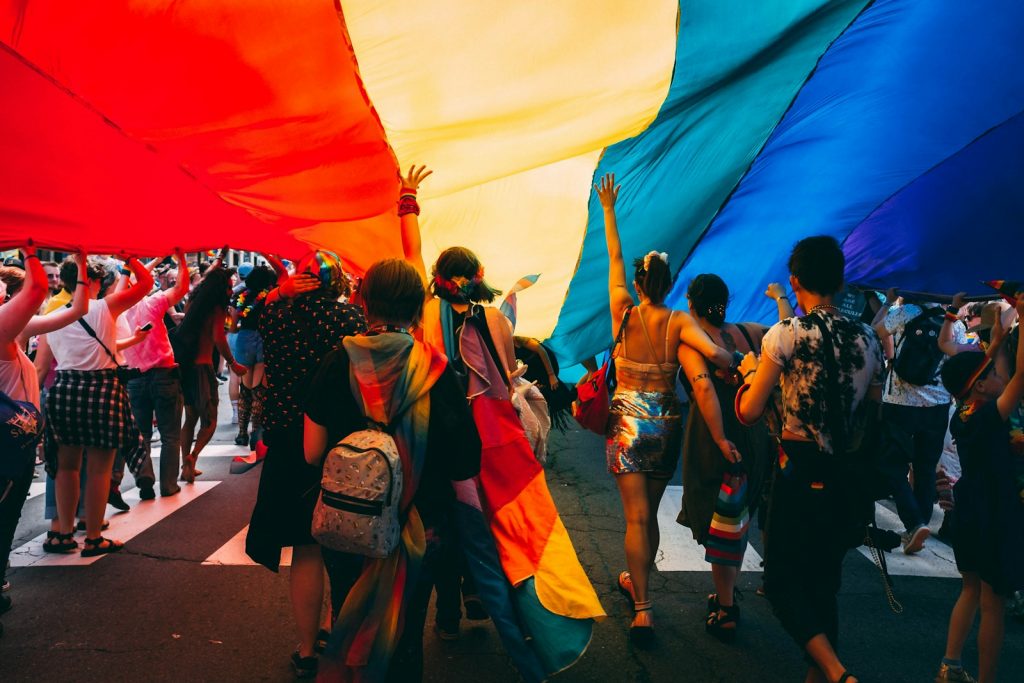 Um grupo de pessoas num evento LGBT caminha sob uma grande e colorida bandeira arco-íris, algumas com as mãos erguidas, usa vários trajes vibrantes. Um grupo de pessoas num evento LGBT caminha sob uma grande e colorida bandeira arco-íris, algumas com as mãos erguidas, usa vários trajes vibrantes.
