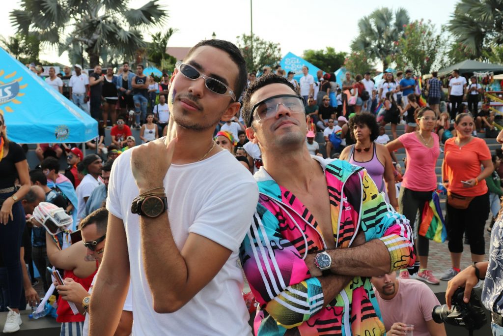 Dois homens, usando roupas elegantes e óculos de sol, posam confiantemente em uma reunião animada ao ar livre, parte da parada vibrante e eventos da cena LGBTI, com uma multidão de pessoas ao fundo. Dois homens, usando roupas elegantes e óculos de sol, posam confiantemente em uma reunião animada ao ar livre, parte da parada vibrante e eventos da cena LGBTI, com uma multidão de pessoas ao fundo.