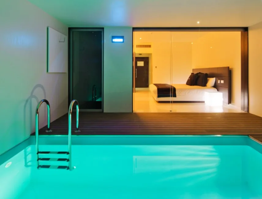 Quarto no Motel H2On com Piscina interior com água iluminada, adjacente a um quarto com uma cama grande, visível através de portas de vidro.