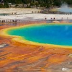 Vista aérea da Grand Prismatic Spring no Parque Nacional de Yellowstone, exibindo cores vívidas em azul, verde, amarelo e laranja que relembram as águas de vários spas. Os visitantes passaram por uma passadeira nas proximidades.