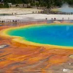 Vista aérea da Grand Prismatic Spring no Parque Nacional de Yellowstone, exibindo cores vívidas em azul, verde, amarelo e laranja que relembram as águas de vários spas. Os visitantes passaram por uma passadeira nas proximidades.