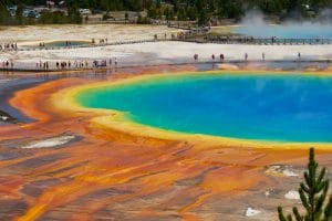 Vista aérea da Grand Prismatic Spring no Parque Nacional de Yellowstone, exibindo cores vívidas em azul, verde, amarelo e laranja que relembram as águas de vários spas. Os visitantes passaram por uma passadeira nas proximidades.
