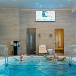 Duas pessoas relaxam numa piscina coberta. A sala possui características, um ecrã que mostra a temperatura e a mudança, plantas em vasos, e uma porta de sauna de madeira perfeita para uma experiência acolhedora de sauna para casal.