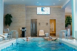 Duas pessoas relaxam numa piscina coberta. A sala possui características, um ecrã que mostra a temperatura e a mudança, plantas em vasos, e uma porta de sauna de madeira perfeita para uma experiência acolhedora de sauna para casal.