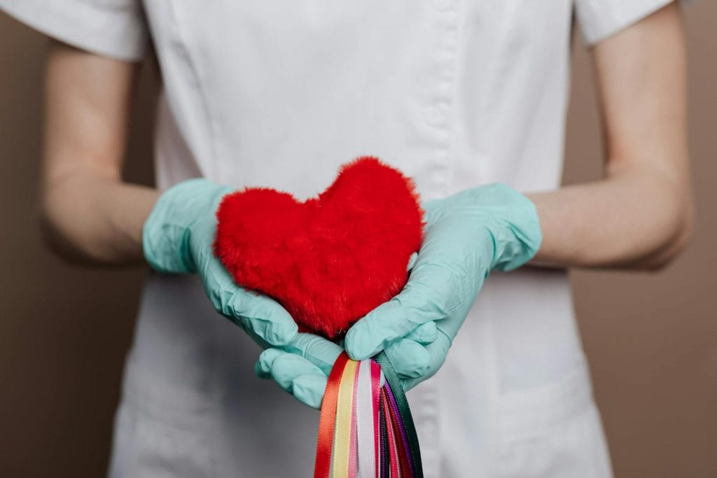 Uma pessoa de bata branca e luvas azuis de segurança um coração de peluche vermelho adornado com fitas coloridas, simbolizando calor e cuidado. Tal como uma sauna beneficia o sistema cardiovascular, esta cena personifica saúde e vitalidade. Uma pessoa de bata branca e luvas azuis de segurança um coração de peluche vermelho adornado com fitas coloridas, simbolizando calor e cuidado. Tal como uma sauna beneficia o sistema cardiovascular, esta cena personifica saúde e vitalidade.