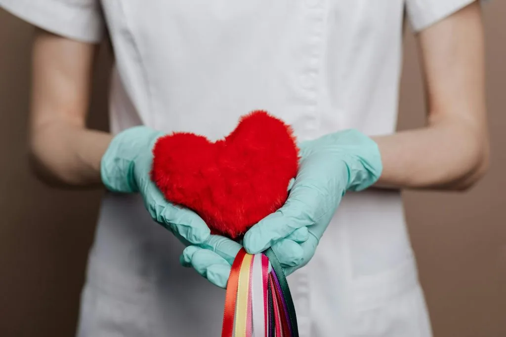 Uma pessoa de bata branca e luvas azuis de segurança um coração de peluche vermelho adornado com fitas coloridas, simbolizando calor e cuidado. Tal como uma sauna beneficia o sistema cardiovascular, esta cena personifica saúde e vitalidade.