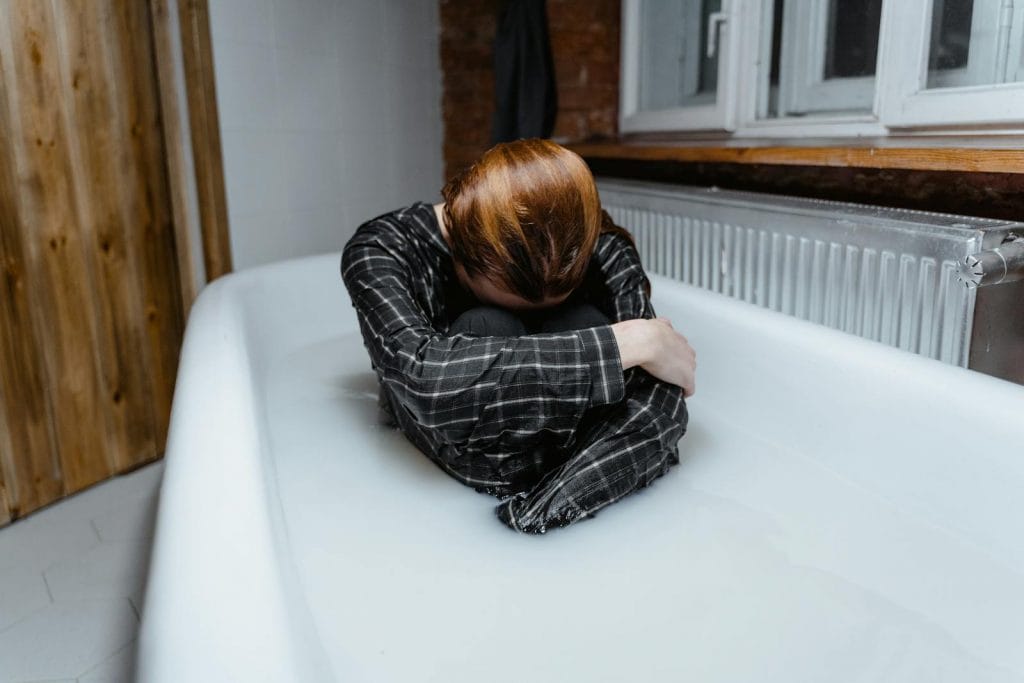 Uma pessoa sentada em uma banheira cheia de água, veste uma camisa axadrezada, com a cabeça ajustada nos joelhos e os braços nas voltas das pernas, personificando a luta silenciosa da depressão na casa de banho tenuemente iluminada — um momento pungente que reflete sobre a saúde mental dentro da comunidade LGBT. Uma pessoa sentada em uma banheira cheia de água, veste uma camisa axadrezada, com a cabeça ajustada nos joelhos e os braços nas voltas das pernas, personificando a luta silenciosa da depressão na casa de banho tenuemente iluminada — um momento pungente que reflete sobre a saúde mental dentro da comunidade LGBT.