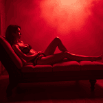 Uma mulher em lingerie preta reclina-se languidamente sobre uma chaise longue, iluminada por uma luz vermelha dramática e envolta numa atmosfera enevoada, evocando o fascínio dos fetiches nos clubes swinguer.