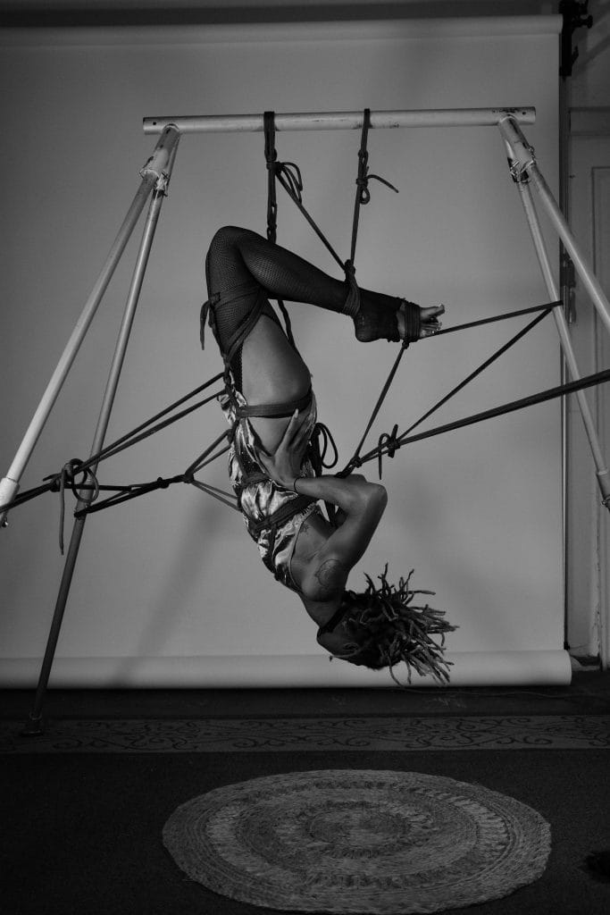 Uma pessoa suspensa de cabeça para baixo num arnês, com cordas presas a uma estrutura metálica, colocada diante de um fundo de estúdio interior. A imagem evidencia a relevância dos cuidados posteriores ao BDSM (aftercare), sublinhando a importância da atenção e do acompanhamento após práticas deste género para o bem-estar físico e emocional das partes envolvidas. Uma pessoa suspensa de cabeça para baixo num arnês, com cordas presas a uma estrutura metálica, colocada diante de um fundo de estúdio interior. A imagem evidencia a relevância dos cuidados posteriores ao BDSM (aftercare), sublinhando a importância da atenção e do acompanhamento após práticas deste género para o bem-estar físico e emocional das partes envolvidas.