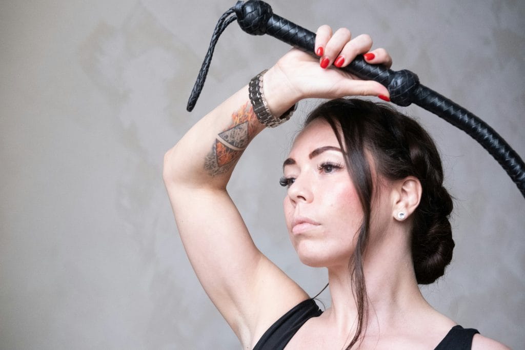 Uma mulher com uma tatuagem no antebraço seguro, acima da cabeça, um chicote de couro preto; mantém o olhar direcionado para a frente, com expressão neutra, encarnando a importância dos cuidados posteriores ao BDSM. Uma mulher com uma tatuagem no antebraço seguro, acima da cabeça, um chicote de couro preto; mantém o olhar direcionado para a frente, com expressão neutra, encarnando a importância dos cuidados posteriores ao BDSM.
