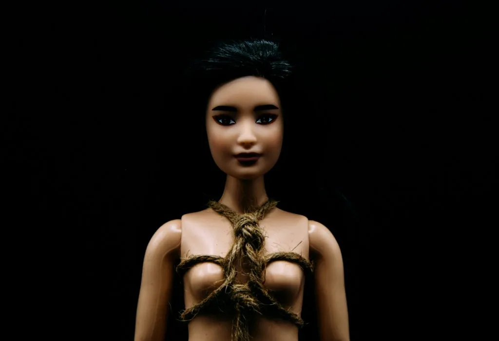 Uma boneca de plástico, com longos cabelos escuros, usando uma corda amarrada em torno do torso, encontra-se posicionada contra um fundo preto, evocando a importância dos cuidados pós-BDSM. Uma boneca de plástico, com longos cabelos escuros, usando uma corda amarrada em torno do torso, encontra-se posicionada contra um fundo preto, evocando a importância dos cuidados pós-BDSM.