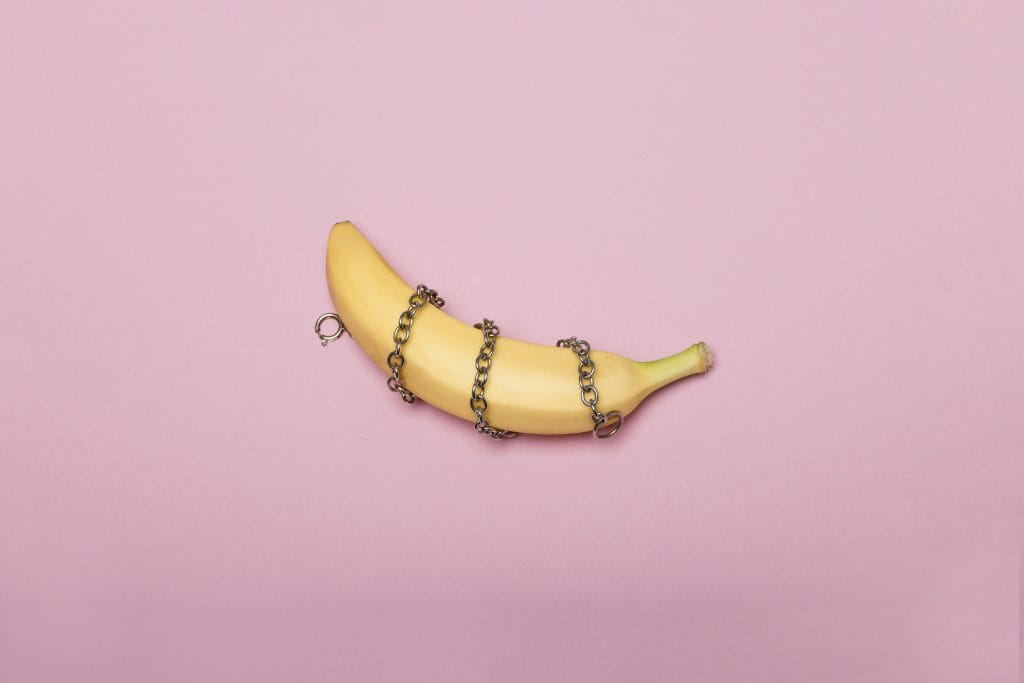 Uma banana amarela com uma corrente metálica enrolada à sua volta, colocada sobre um fundo cor-de-rosa claro, simboliza o cuidado pós BDSM de forma lúdica e visualmente marcante. Uma banana amarela com uma corrente metálica enrolada à sua volta, colocada sobre um fundo cor-de-rosa claro, simboliza o cuidado pós BDSM de forma lúdica e visualmente marcante.
