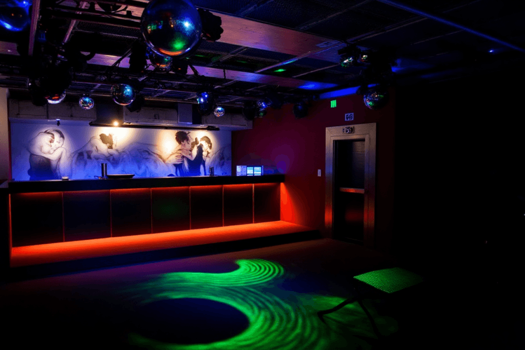 A discoteca Trumps em Lisboa. Uma discoteca pouco iluminada com um bar em tons vermelhos, bolas de espelhos no teto, luzes coloridas no chão e um mural de figuras a dançar atrás do bar. A discoteca Trumps em Lisboa. Uma discoteca pouco iluminada com um bar em tons vermelhos, bolas de espelhos no teto, luzes coloridas no chão e um mural de figuras a dançar atrás do bar.