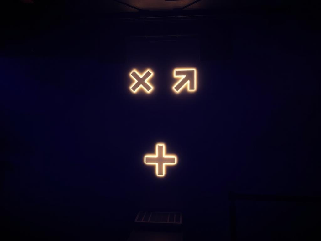 Os ícones estão em uma parede escura: um "X", uma seta para cima e direita, e um sinal de mais, evocando uma linguagem inclusiva. Os ícones estão em uma parede escura: um "X", uma seta para cima e direita, e um sinal de mais, evocando uma linguagem inclusiva.