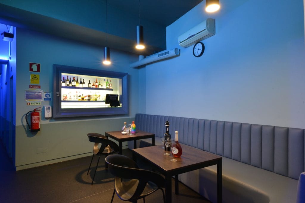 Interior moderno do Snack-Bar da SaunApolo 56, com duas mesas, bancos estofados cinzentos, luzes pendentes e expositores de bebidas atrás de vidro. Interior moderno do Snack-Bar da SaunApolo 56, com duas mesas, bancos estofados cinzentos, luzes pendentes e expositores de bebidas atrás de vidro.