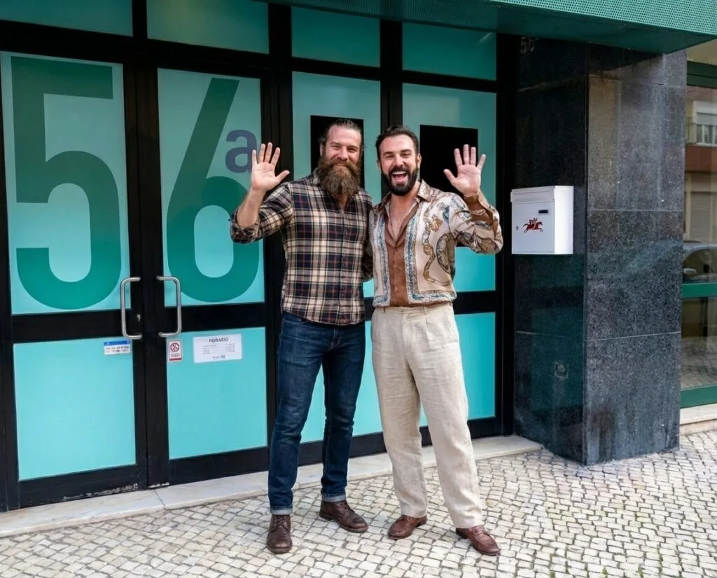 Dois homens sorriem e acenam diante da porta de vidro com o número "56a", recebendo visitantes na SaunApolo 56. Dois homens sorriem e acenam diante da porta de vidro com o número "56a", recebendo visitantes na SaunApolo 56.