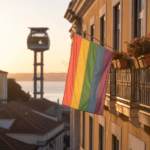 Bandeira arco-íris na varanda com floreiras, sobre telhados de Lisboa e torre do teleférico ao pôr do sol; ambiente acolhedor.